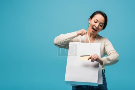 Excited asian customer holding shopping bags and credit card isolated on blue 