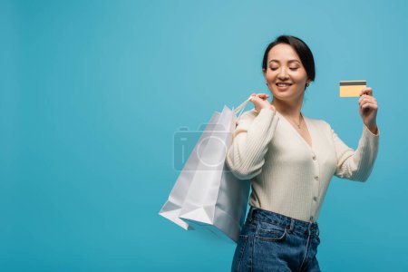 Happy asian woman holding credit card and purchases isolated on blue 