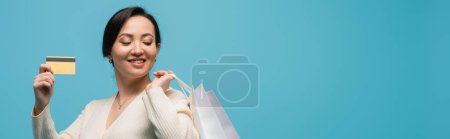 Positive asian shopaholic holding shopping bags and credit card isolated on blue, banner 
