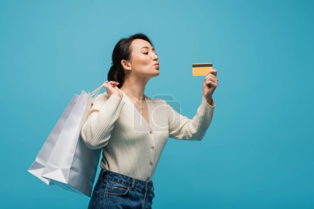 Young asian model pouting lips while holding credit card and shopping bags isolated on blue 