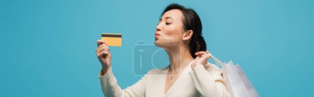 Asian woman holding shopping bags and pouting lips while looking at credit card isolated on blue, banner 