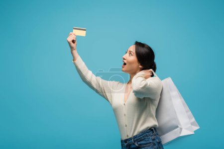 Amazed asian woman holding chopping bags and looking at credit card isolated on blue 