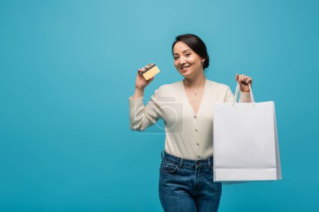 Young asian customer holding shopping bags and credit card isolated on blue 