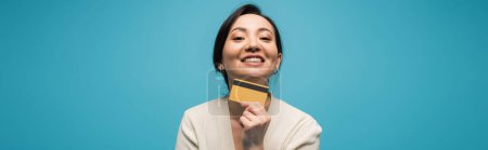 Smiling asian model holding credit card isolated on blue, banner 