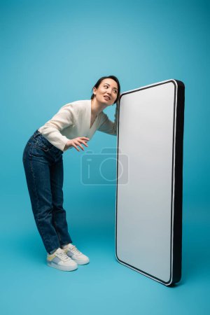 Full length of young asian woman standing near smartphone mockup on blue background