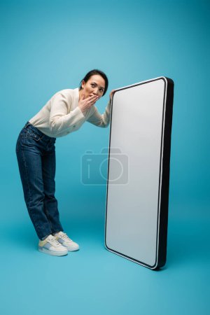 Shocked asian woman covering mouth near big smartphone model on blue background