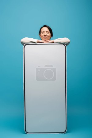 Pretty brunette woman standing near big cellphone model on blue background