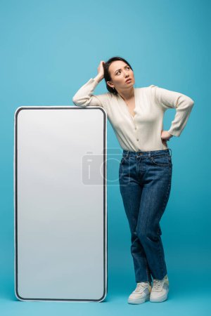 Full length of pensive asian woman standing near big smartphone mockup on blue background