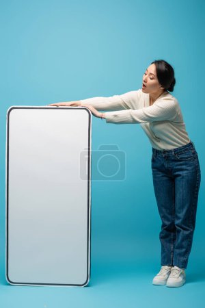 Shocked asian woman in jeans and cardigan standing near big smartphone model on blue background