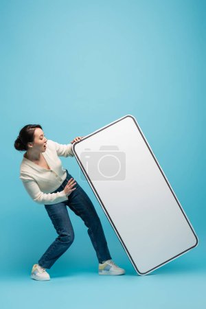 Shocked asian woman looking at big cellphone model on blue background
