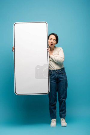 Shocked asian woman holding big smartphone model on blue background