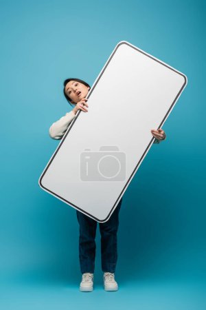 Confused asian woman holding big smartphone model on blue background