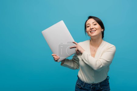 Positive asian model in cardigan holding laptop and looking at camera isolated on blue 