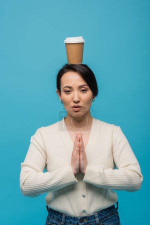 Asian woman with coffee to go on head doing praying hands gesture isolated on blue 