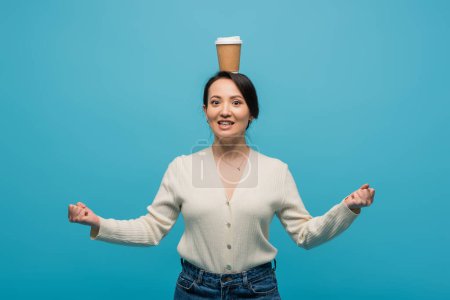 Excited asian woman with paper cup on head showing yes gesture isolated on blue 
