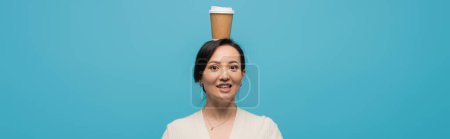 Smiling asian woman holding coffee to go on head isolated on blue, banner 
