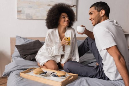 Photo for Happy african american couple sitting with coffee and orange juice near tray with pancakes in bedroom - Royalty Free Image