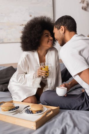 Photo for Young african american couple with coffee and orange juice smiling near tray with pancakes and berries on bed - Royalty Free Image