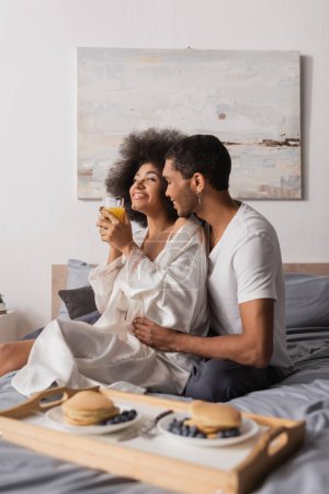 Téléchargez les photos : Heureuse femme afro-américaine en robe de soie tenant du jus d'orange pendant le petit déjeuner avec petit ami au lit - en image libre de droit
