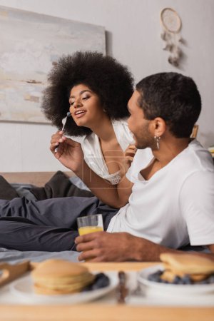 Photo for Young african american man holding orange juice and feeding sexy girlfriend with berry during breakfast - Royalty Free Image