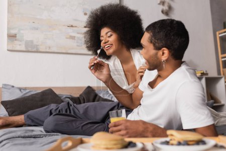 Photo for African american man holding fork with blueberry near cheerful girlfriend during breakfast in bedroom - Royalty Free Image