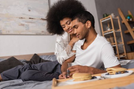 Photo for Pretty african american woman smiling near young boyfriend and delicious pancakes on blurred foreground in bedroom - Royalty Free Image