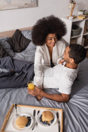 Photo for High angle view of african american woman smiling near young boyfriend and delicious pancakes with blueberries on bed - Royalty Free Image