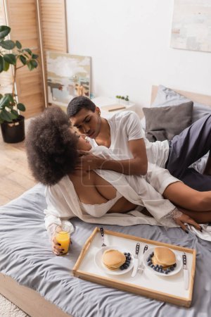 Téléchargez les photos : Vue grand angle de l'homme afro-américain toucher petite amie sexy tenant jus d'orange près de crêpes avec des baies sur le lit - en image libre de droit