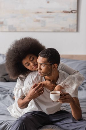 Photo for Positive african american woman embracing young boyfriend sitting with coffee cup on bed - Royalty Free Image