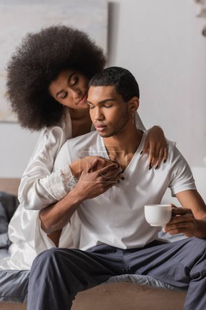 Photo for Young african american man holding coffee cup while sitting with closed eyes near sexy girlfriend hugging him in bedroom - Royalty Free Image