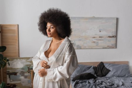 curly african american woman in white satin robe standing and looking away in bedroom