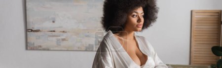 brunette and curly african american woman in white silk robe standing and looking away, banner