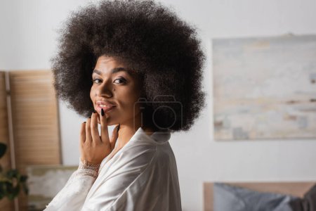 Photo for Seductive african american woman in white silk robe touching lip and smiling at camera at home - Royalty Free Image