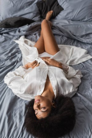high angle view of passionate african american woman in white robe and lingerie lying on grey bedding