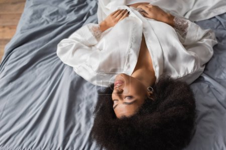 vista de ángulo alto de morena mujer afroamericana con los ojos cerrados acostado en bata blanca sobre ropa de cama gris
