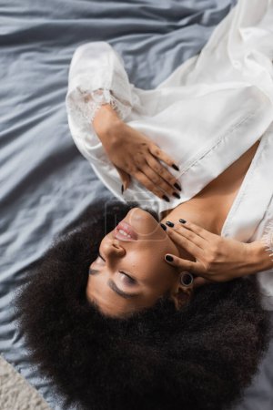 top view of sexy african american woman in white silk robe lying with closed eyes on grey bedding
