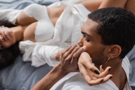 jeune homme afro-américain baisant la main d'une femme passionnée en lingerie blanche couchée sur fond flou