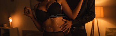 Téléchargez les photos : Vue recadrée de l'homme afro-américain étreignant femme sexy en soutien-gorge noir dans la chambre noire avec éclairage, bannière - en image libre de droit