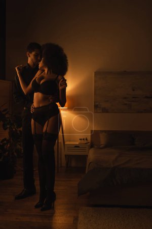 Photo for Full length of african american woman in black underwear standing near young man in dark room with lighting - Royalty Free Image