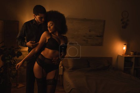 junger Mann berührt sexy afrikanisch-amerikanische Freundin in schwarzen Dessous im dunklen Schlafzimmer mit Beleuchtung