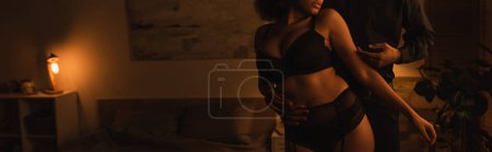 Téléchargez les photos : Vue recadrée de femme afro-américaine passionnée en sous-vêtements près du jeune petit ami dans la chambre la nuit, bannière - en image libre de droit