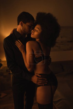 Foto de Seductive african american woman in sexy lingerie embracing with young boyfriend at home at night - Imagen libre de derechos