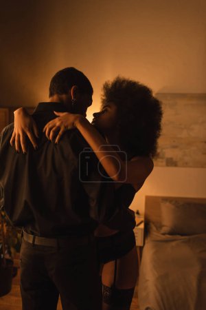 Téléchargez les photos : Femme bouclée et sexy étreignant petit ami afro-américain en chemise noire dans la chambre noire avec éclairage - en image libre de droit