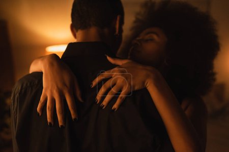 Téléchargez les photos : Séduisante femme afro-américaine embrassant l'homme en chemise noire dans une pièce sombre avec éclairage - en image libre de droit