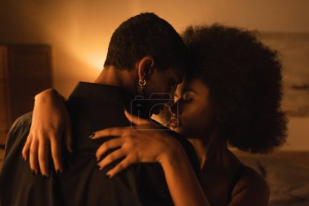 Téléchargez les photos : Jeune femme afro-américaine avec les cheveux bouclés étreignant jeune petit ami en chemise noire dans la chambre sombre - en image libre de droit