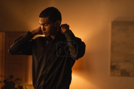 Téléchargez les photos : African american man adjusting black shirt in evening at home - en image libre de droit