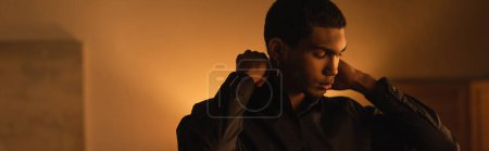 young african american man adjusting collar of black shirt in evening at home, banner