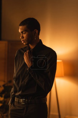Téléchargez les photos : Brunette homme afro-américain en chemise noire debout avec la main dans la poche de pantalon dans la chambre avec lampe lumineuse - en image libre de droit