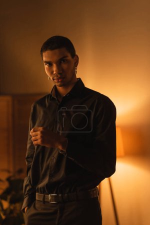 african american guy in black shirt holding hand in pocket of pants and looking at camera in room with lighting
