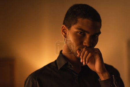 Téléchargez les photos : Pensive african american man in black shirt holding hand near mouth while looking at camera in evening at home - en image libre de droit